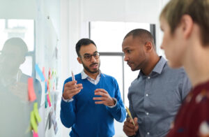 A photo of two male workers and one female worker having a working conversation at a whiteboard.