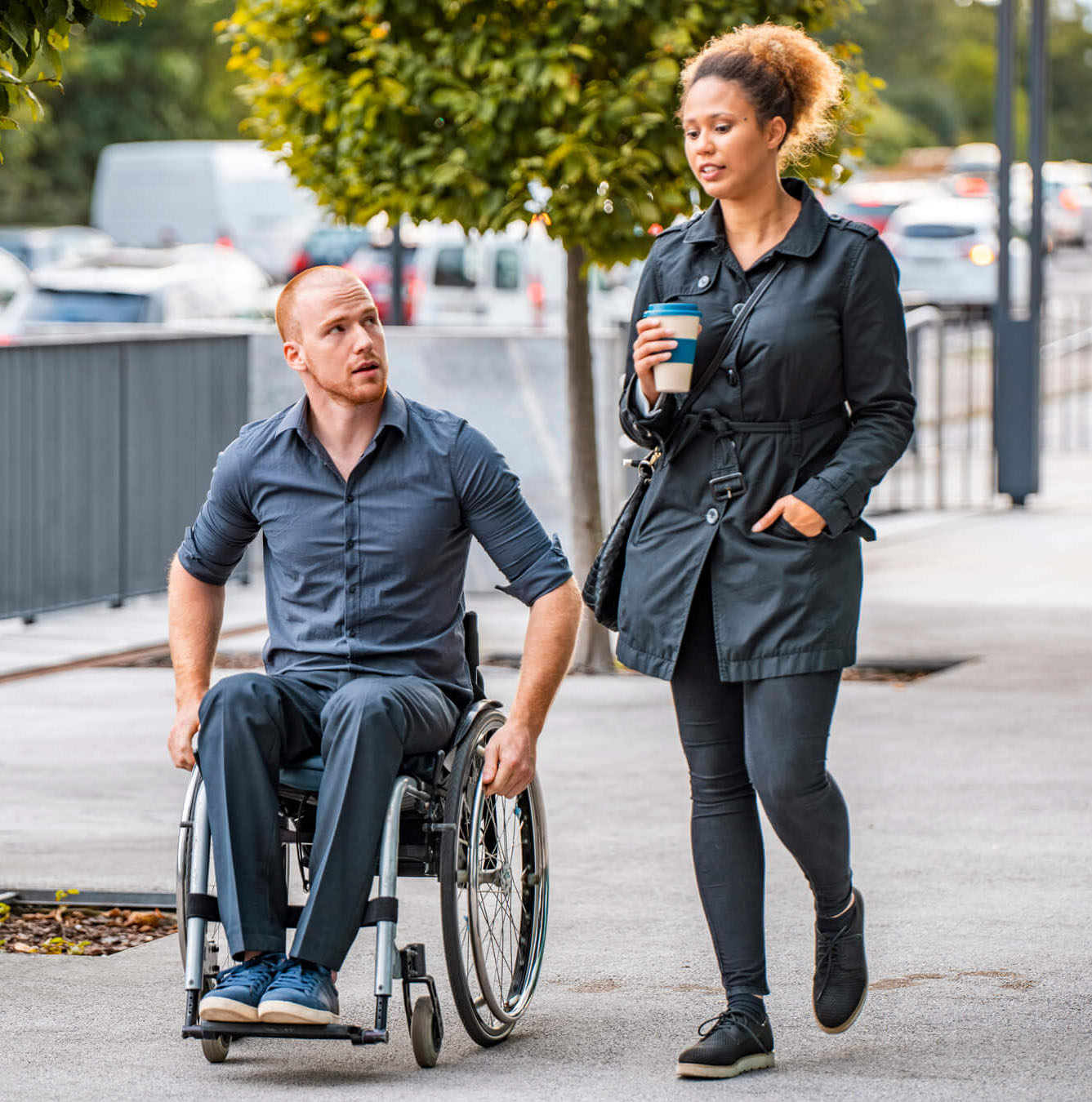 A young man in a wheelchair is strolling alongside a woman holding a coffee cup on a street sidewalk