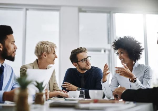 A group of office workers conversing at a conference table, listening intently to the woman speaking.