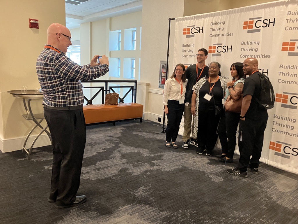 Man taking a photo of group in front of a sign at the CSH Summit in 2023